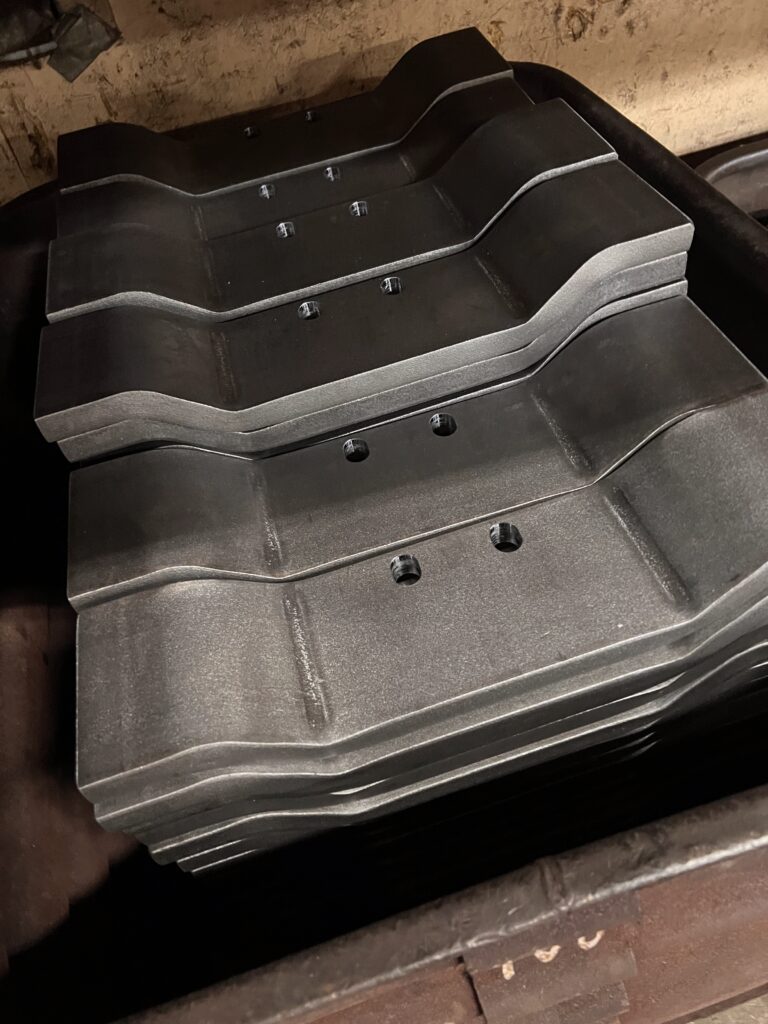 Stack of precision-fabricated metal components with contoured profiles and drilled mounting holes, finished in a dark coating and arranged in a workshop setting.