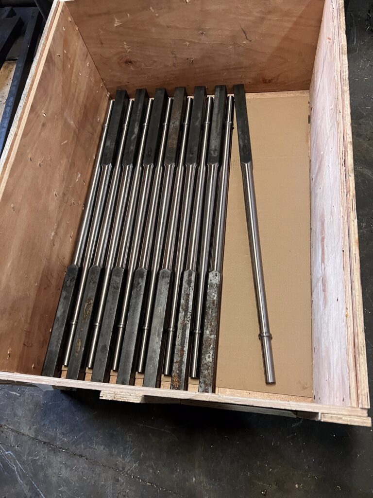 Fabricated architectural metal elements—precision-machined steel rods with shaped ends—neatly arranged inside a wooden crate for storage or transport.