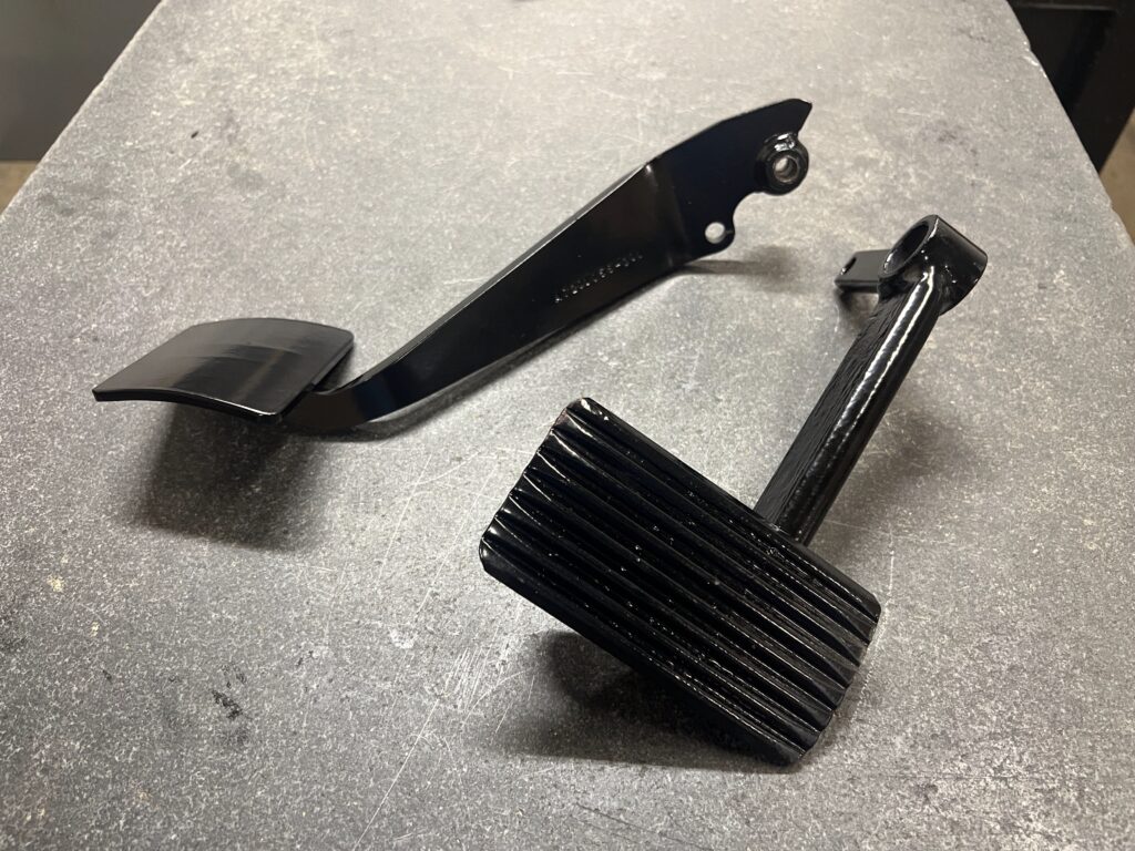 Black metal automotive pedal assembly components, including a ribbed pedal pad and a curved mounting bracket, displayed on a gray textured surface.
